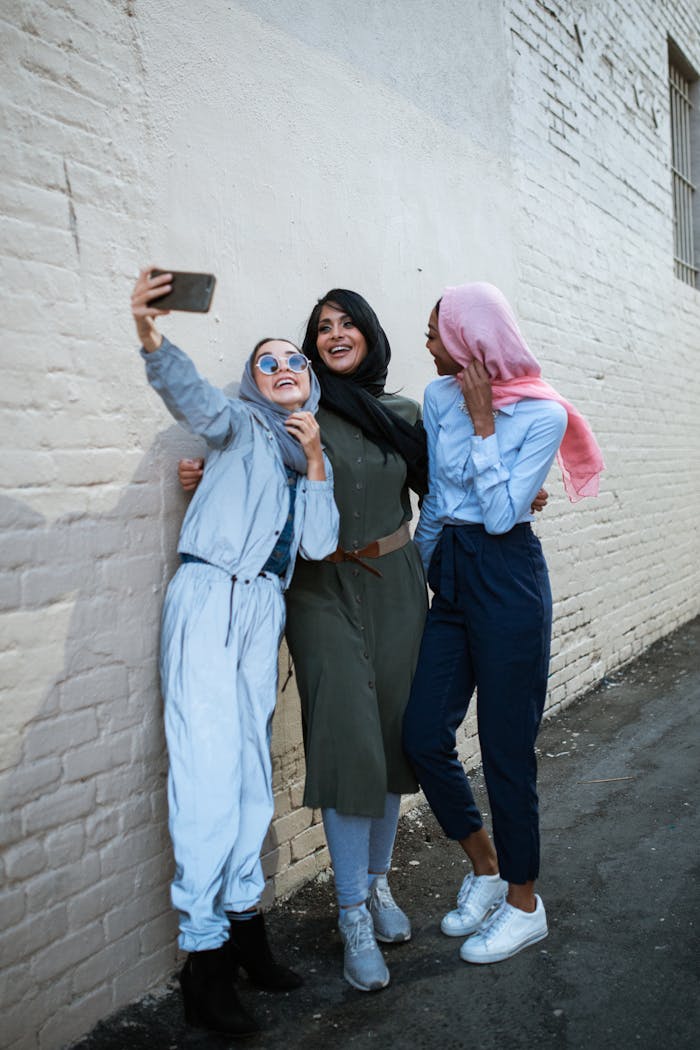 Three diverse friends in hijabs joyfully taking a selfie against a brick wall.